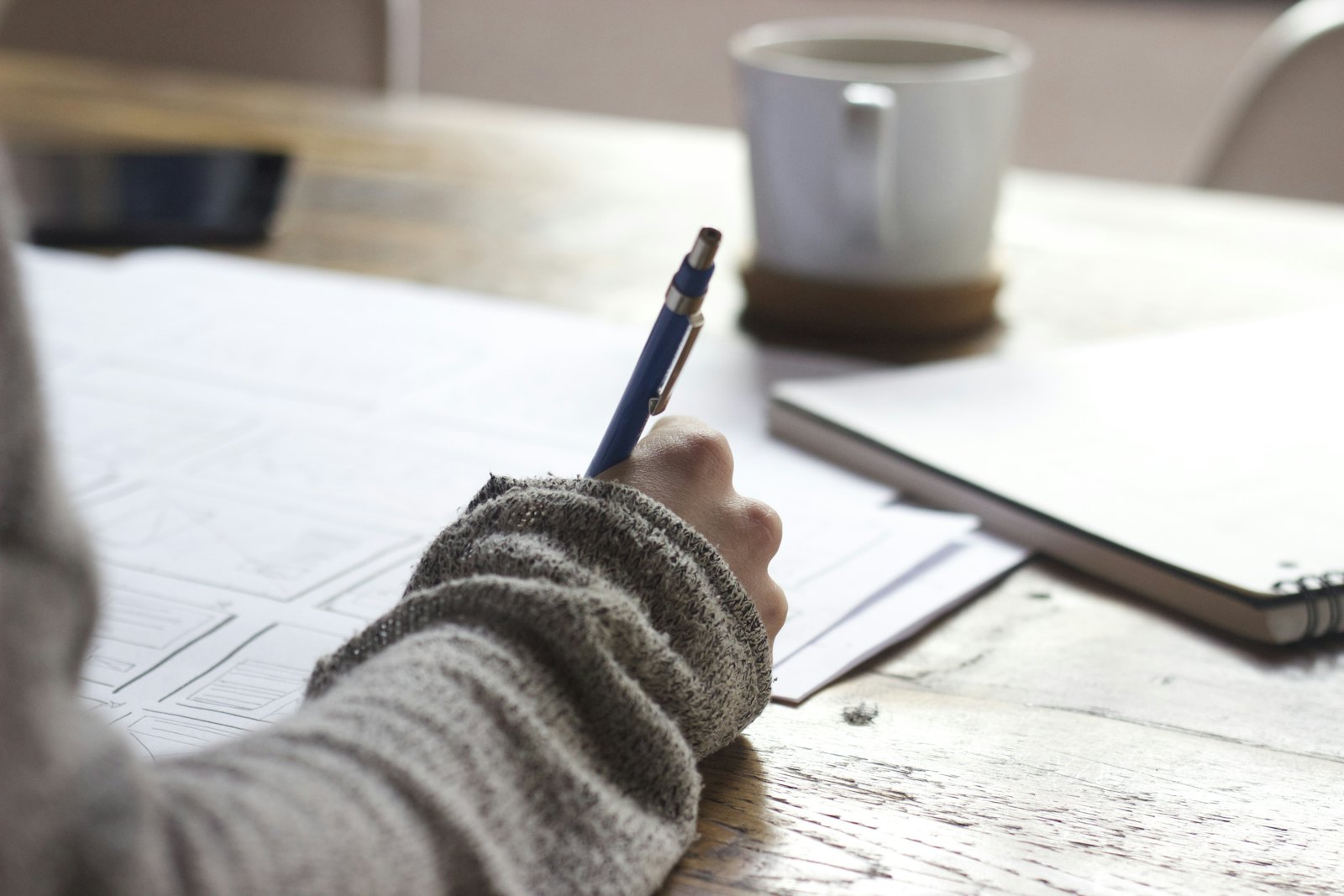 A person sketching on paper with a pen, beside a notebook and a mug.